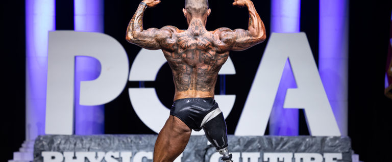 A male bodybuilder poses on stage with his back to the camera, flexing his arms in a double biceps pose.