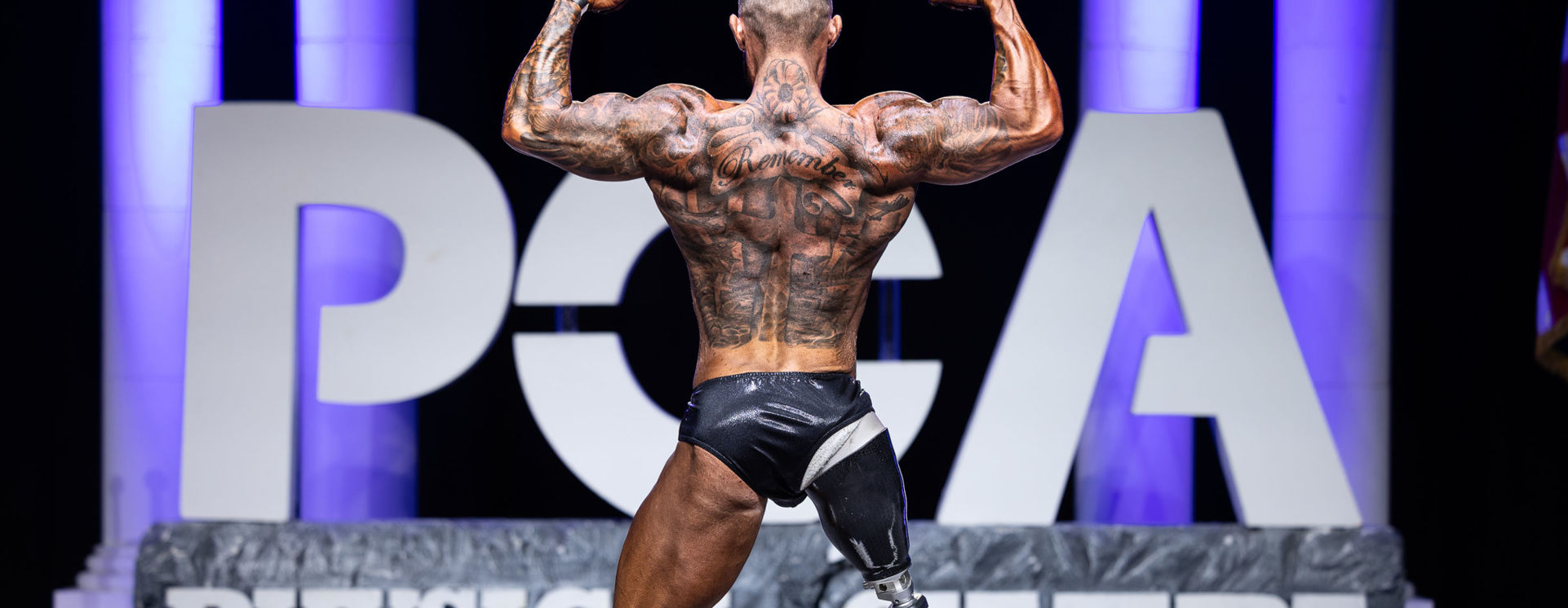 A male bodybuilder poses on stage with his back to the camera, flexing his arms in a double biceps pose.
